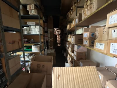 This image depicts a storage or warehouse environment, likely in Campana, Argentina, given the text "HECHO EN CHINA" and product packaging written in Spanish. The scene shows narrow aisles lined with shelves filled with cardboard boxes of varying sizes. The boxes are densely packed, suggesting an efficient use of space. Some boxes are labelled with names like "TRIPTO" and "DAISA." The lighting is dim, making it difficult to discern finer details. There are no people visible in the immediate scene. The overall impression is of a busy storage area containing a variety of products, potentially related to electrical components or manufacturing materials.