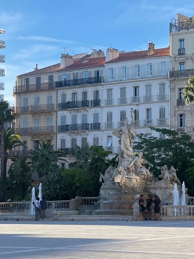 This is a daytime, eye-level shot of the Place Puget in Toulon, France, featuring the iconic Fontaine de la Pyramide. The fountain, a large stone sculpture depicting mythological figures and a central female statue holding a torch, dominates the center of the frame. Water cascades from multiple tiers of the fountain. In the foreground, a paved public square is visible, with several pigeons scattered about. To the left of the fountain, two men stand near a smaller water feature. To the right, three young men in casual attire are gathered, observing the fountain. Behind the fountain and to the left, a row of multi-story buildings with ornate balconies and shuttered windows line the square. Palm trees and other greenery are interspersed between the buildings and around the fountain, adding a lush contrast to the urban architecture. The sky is clear and blue, indicating fair weather.