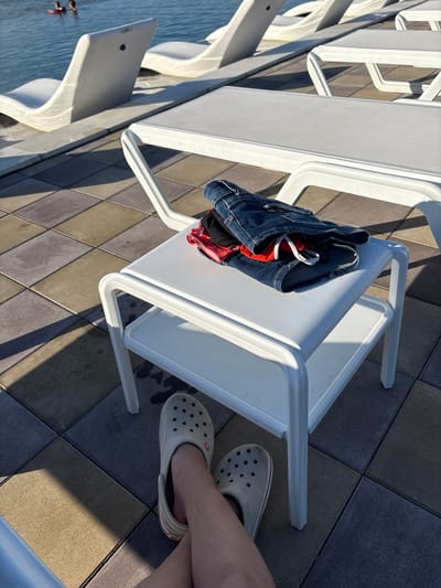The image shows a sunny outdoor scene, likely near a body of water. In the foreground, a person's legs are visible, wearing light-colored Croc-style shoes. The feet are crossed, and the person is wearing denim shorts. To the right, a white side table holds a pair of folded denim shorts and some red items underneath. The table has two tiers. In the background, white lounge chairs are arranged near a pool or lake, with two people in the water. The ground is paved with square tiles in various shades of brown, gray, and beige. The scene appears to be in Kamiani Potoky, Ukraine, a location known for its natural beauty and recreational areas along the Dnieper River.