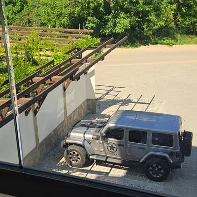 A silver Jeep Wrangler is parked on a cobblestone surface next to a white wall and a wooden pergola structure. The Jeep has a spare tire mounted on the rear and star decals on the side. The environment is outdoors, with green trees and foliage visible in the background, and a paved road adjacent to the parking area. The scene appears to be in Krasnovo, Bulgaria, indicated by the context provided. The lighting suggests it is daytime, with shadows cast by the pergola and the vehicle on the ground. No people are visible in the image.