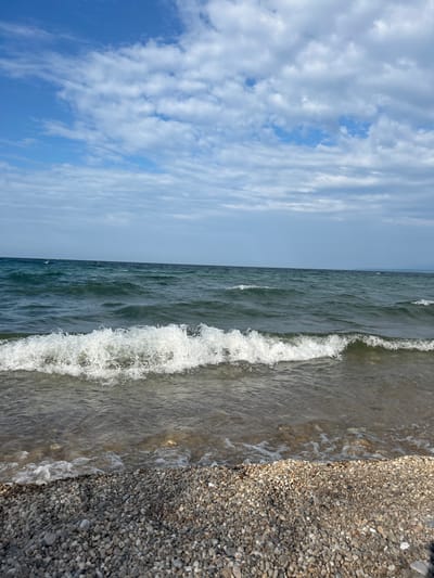 The image depicts a natural scene at the shore of a body of water. In the foreground, a pebbled beach is visible, with small, rounded stones in shades of gray and beige. The water's edge meets the pebbles, with small waves lapping at the shore, creating white foam and ripples. Further out, the water appears dark green and choppy, with larger waves rolling in. The horizon line is visible in the distance, separating the water from the sky. The sky is a clear blue with scattered white, wispy clouds. There are no people or man-made structures present in the image. The location is identified as being in the city of Tolbazikha, Russia, implying this is a natural landscape within that area.