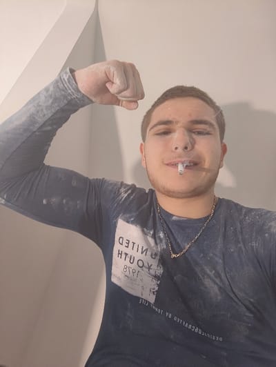 A male individual, appearing to be in his late teens or early twenties, is depicted in an indoor setting. He has short brown hair, light stubble, and a small, dark piercing or mark on the bridge of his nose. He faces the camera directly with a slight smile, a lit cigarette held between his lips. He is wearing a dark blue, long-sleeved t-shirt with a reversed white graphic print on the chest that reads "UNITED YOUTH 1978" and "DESIGNED BY TOMMY LIFE." A thin gold-colored chain necklace is visible around his neck. His left arm is raised and bent, forming a bicep flex with a clenched fist. His face, hair, left arm, and clothing are covered in a fine white powdery substance, consistent with plaster or drywall dust. The background consists of plain, light-colored walls, with an interior corner visible behind his raised arm. The indoor environment suggests a space under construction or renovation. The location in Deerlijk, Belgium, cannot be determined from the visual information provided in the image.