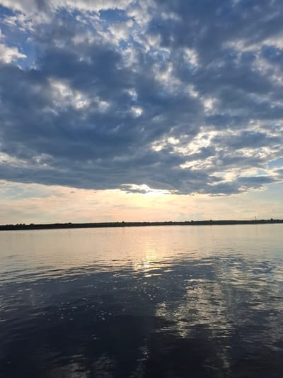 The image depicts a scenic view of clouds and a body of water, likely a river, with the sun setting or rising. The sky is filled with dynamic clouds, ranging from dark, stormy gray to lighter, wispy white formations. The sun's rays are breaking through the clouds, creating a bright glow that illuminates the scene.
The body of water occupies the right side of the frame and reflects the sky's colors. The surface of the water is rippled, showing the effect of a gentle breeze. A thin strip of land with dark, silhouetted trees or vegetation is visible along the horizon, separating the water from the sky. In the distance, a slender tower or pole stands against the sky.
The overall mood of the image is peaceful and serene, with a sense of natural beauty. The time of day is indicated by the warm, golden light of the sun, suggesting either sunrise or sunset. The weather appears to be partly cloudy, with dramatic cloud formations. The location is suggested to be Введеновка, Russia, which aligns with the natural landscape shown. No people or man-made objects other than the distant tower are clearly visible in the main frame.