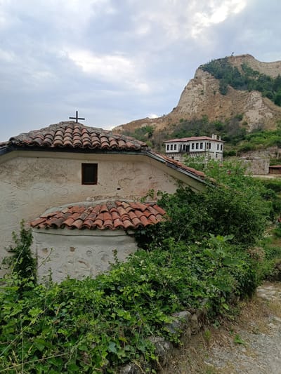The image captures an exterior view of a small, old stone church with a red tiled roof, topped by a black cross. The church's walls are textured, with a subtle decorative pattern, and a small, dark-paned window is visible. Overgrown green ivy and other plants climb the walls and surround the structure, creating a lush, somewhat wild atmosphere.

In the background, a prominent, steep, sandy-colored hill rises, dotted with sparse trees and vegetation. At the foot of the hill, a larger, white building with a red roof and dark windows stands, suggesting a historical settlement. The setting appears to be a warm day, possibly late afternoon or early morning, given the soft light. The sky is cloudy, with patches of bright sky breaking through.

The scene evokes a sense of history and tranquility. The combination of the ancient church, the natural landscape, and the traditional architecture suggests a location with deep historical roots. The lush greenery hints at a temperate climate. The image is taken from a slightly low angle, emphasizing the church in the foreground and the imposing hill behind it. The location is identified as Melnik, Bulgaria, a town known for its historical architecture and unique geological formations. No text is visible in the image.
