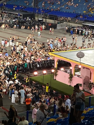 The image depicts a large crowd of people gathered in an outdoor stadium setting at night. The scene appears to be some kind of event or concert, with a stage or performance area visible in the background, characterized by lights and structures. People are standing and milling about in a large open area, with rows of blue stadium seating visible on the left side of the image. The overall atmosphere suggests a lively gathering.
The subjects are primarily people, dressed in casual summer attire such as t-shirts, shorts, and dresses. There are many individuals and small groups interacting within the crowd. Objects visible include the stage structure, lighting equipment, a satellite dish mounted on a pole, and the stadium seating. The setting is clearly a stadium, likely in an urban or suburban area, given the surrounding infrastructure.
The activity is people attending an event, likely a music performance or festival, as indicated by the stage and the crowd's engagement. The time of day is clearly night, with artificial lighting illuminating the scene. The weather appears to be warm, judging by the clothing. There are no obvious location cues specific to Arnhem, Netherlands, beyond the provided context. Emotions are not clearly discernible due to the distance and low resolution of many individuals, but the overall impression is one of social gathering and possibly enjoyment. No visible text is present in the image.