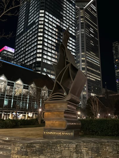 A nighttime view showcasing a sculpture of upright books with a metal structure extending above them, possibly representing pages or wings. The sculpture bears the name "Ueainius Koy" on one of the books. A modern skyscraper, brightly lit with numerous windows, looms in the background. A building with a distinctive gable roof is also visible near the base of the book sculpture. The scene is illuminated by city lights.