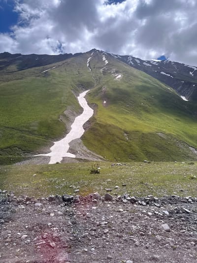 A mountain slope in Almasiani, Georgia, is covered in green grass with patches of snow. A winding strip of snow descends the mountain. The sky above is cloudy. The foreground is a rocky, gravelly area.