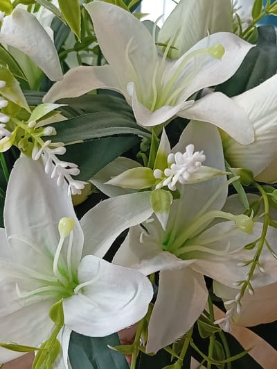 A close-up, overhead shot of an artificial white lily arrangement is presented. The focus is on the intricate details of the lilies, their petals, stamens, and pistils, alongside lush green foliage and smaller white decorative flowers. The background is softly blurred, offering no discernible environmental context or geographical markers for Kerben, Kyrgyzstan. No individuals or actions are depicted in the image.