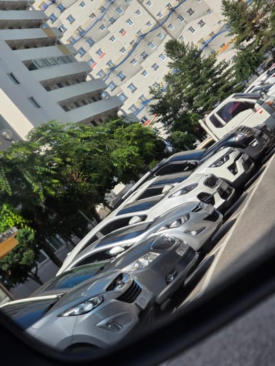 The image shows a street in Eumseong-eup, South Korea, with several cars parked in a row. The cars are mostly white and silver, and they appear to be modern models. In the background, there is a tall apartment building with many windows. Trees and foliage are visible on the right side of the image. The scene is bathed in sunlight, suggesting it is daytime. The perspective is from inside a vehicle, possibly looking out of a side mirror. No people are clearly visible in the image. The overall impression is of a typical urban street scene.