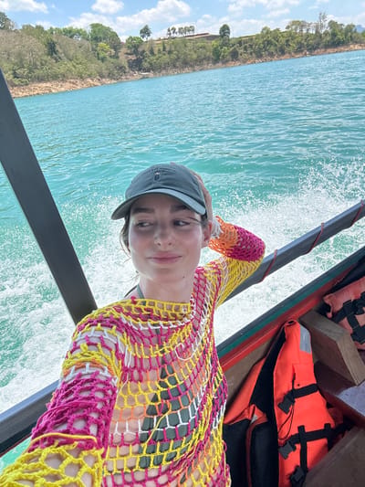 A medium shot captures a woman on a boat in Ban Ta Khun, Thailand. She wears a gray cap and a colorful, netted top in shades of pink, yellow, white, and orange. The boat moves through turquoise water, generating spray. Behind her, a shoreline with trees is visible. In the boat, life jackets are stowed.