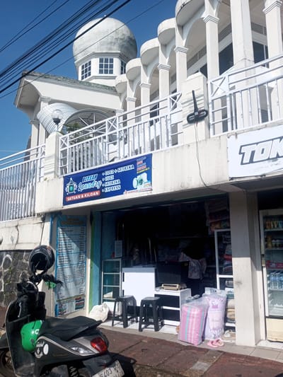 A street-level view shows the "Berkah Laundry" storefront in an unidentified city. The laundry is located on the ground floor of a white building with a rounded dome and arched columns. A blue sign with the laundry's name hangs above the entrance, advertising washing, ironing, and express services. A smaller sign to the side lists prices. Two pink and white striped laundry bags sit on the sidewalk along with a pair of pink flip-flops. A black and grey scooter with a green helmet and a black helmet parked on it is in the foreground. Next to the laundry is another storefront with a "TOK" sign. A person in a patterned shirt is visible inside the laundry. The sky is clear and blue. Overhead power lines are also visible. The location in Nhot Chang, Laos cannot be verified from the provided image.