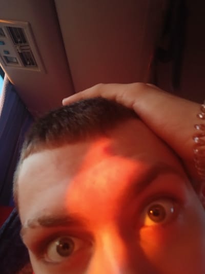 A close-up selfie shows a person's face illuminated by an orange light, casting a warm glow on their features. The person has a short haircut and light skin. Their eyes are wide, with visible brown irises. The person's hand is visible on the right side of the frame, resting against their head. Behind them, a panel with vent-like openings is visible. The image does not contain enough information to confirm it was taken in Ustinovo, Russia.