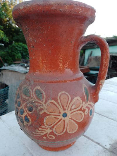 The central subject of the image is an old, reddish-brown terracotta or clay pitcher, resting on a white, slatted table or surface. The pitcher features a bulbous body, a narrower neck, and a thick, slightly flared rim. A sturdy, C-shaped handle is attached from the upper body to the neck.
The surface of the pitcher appears weathered and rustic, with a coarse texture and numerous specks of dirt and dust, particularly visible on the rim and around the patterned areas. A delicate cobweb is draped across the inner curve of the handle, indicating its age and outdoor placement. The primary color of the pitcher is a warm, earthy red-orange, with what appears to be a worn or imperfect glaze that gives it a slight sheen in some areas.
Around the lower half of the pitcher, there are hand-painted floral and organic patterns. These designs feature stylized flowers with lighter orange or beige petals and dark blue centers, interspersed with dark blue and green leaf-like or abstract shapes, all outlined in a lighter, yellowish hue. A thin, horizontal line in greenish-blue runs along the base of the decorated section.
The background is softly blurred, revealing an outdoor setting during the daytime. Green foliage, likely trees or bushes, is visible, along with vague outlines of what appear to be simple, rustic structures or fences. The bright sky is visible in some areas. The pitcher is stationary, and there are no people or discernible activity taking place. No text is visible in the image. The scene suggests a quiet, informal outdoor environment, possibly a garden or backyard.