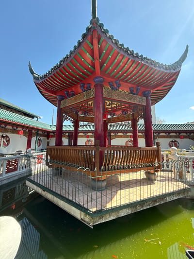 The image, correctly oriented, depicts a vibrant Chinese-style pavilion or section of a temple situated next to a body of water, all under a clear blue sky. The central structure features a distinctive, multi-tiered roof with gracefully curved eaves, adorned with red and green tiles. The underside of the eaves is intricately decorated with traditional patterns and what appear to be small, dark-colored ornaments.
The pavilion is supported by sturdy red wooden columns, some embellished with gold-colored details and dark purple, sculpted brackets. Inside the structure, detailed carved wooden lattices and panels are visible, along with a long wooden bench or railing.
Adjacent to the pavilion is a calm body of water, likely a pond or canal, which has a distinct green hue. Several small orange fish can be seen swimming beneath its surface. A metal railing separates the tiled platform or walkway from the water. Bright sunlight illuminates the scene, casting strong shadows of the railing onto the geometric pattern of the tiles.
In the background, other buildings designed in a similar traditional Chinese architectural style are visible, showcasing complementary rooflines and white walls adorned with round decorative motifs that feature figures and patterns.
The scene takes place during the daytime, under bright sunlight and a clear blue sky, indicating pleasant weather. No people are visible, emphasizing the architectural and natural elements. The presence of this distinctly Chinese architecture within Hainburg an der Donau, Austria, suggests a specific cultural garden or themed area. No clearly legible text is visible in the image.