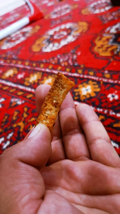 A person's hand is holding a single, golden-brown crouton against a backdrop of a red, patterned fabric. The crouton is elongated and has a porous texture, suggesting it is made from toasted bread. The hand is a light to medium skin tone, and the fingers are gently cupping the crouton. The fabric behind the hand features intricate geometric and floral designs in shades of red, orange, yellow, and black, resembling a traditional rug or tapestry. In the upper left corner of the image, a portion of a white and red object, possibly a pen or marker, is visible. The lighting suggests an indoor setting with a light source from the top left. The scene appears to be a close-up, personal moment, perhaps of someone about to eat the crouton or examining it. The location is not specified as being in the city of Onaldy, Turkmenistan.
