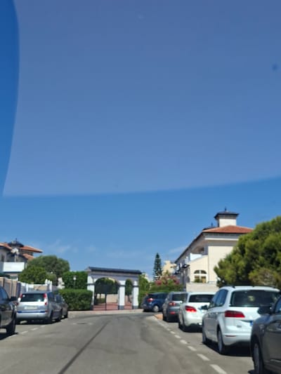 The image shows a street in Cabo Cervera-Playa La Mata, Spain, lined with parked cars. The scene appears to be set during the daytime, with bright sunlight casting shadows. The sky is a clear, vibrant blue.

The foreground is dominated by the reflection of the sky on what appears to be a windshield or window. Beyond this reflection, a street is visible with several cars parked along the side. The cars are of various makes and colors, including white SUVs, a dark-colored sedan, and a darker vehicle partially visible at the bottom.

To the right of the street, there are buildings and lush green trees. The architecture suggests a residential or commercial area with houses and perhaps shops. Some buildings have terracotta-tiled roofs. There is also a glimpse of what looks like a white building with arched features and columns, possibly an entrance to a property or a public space.

The overall impression is a peaceful, sunny day in a coastal town. There are no people visible in the image, and the focus is on the static elements of the scene. The image is oriented vertically, with the sky occupying the left half and the street scene the right. There is no visible text in the image.