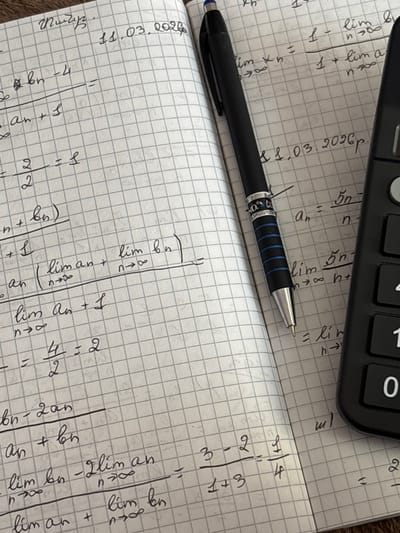 This image shows a close-up of a notebook with handwritten mathematical equations. A black pen is positioned on the open pages of the notebook, and part of a black calculator is visible on the right side. The notebook has a grid pattern on its pages. The handwriting includes mathematical notation such as fractions, limits, and variables like 'a' and 'b'. Some of the equations appear to be calculations involving sequences and limits. A date, "11.03.2026", is also written on the page. The setting is a desk or table, suggested by the texture of the surface beneath the notebook. The image does not contain any information about the city of Jrarat, Armenia.