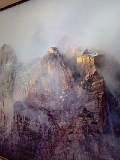 A mountain range shrouded in mist and fog dominates the scene. Jagged peaks rise through the clouds, revealing sections of exposed rock and snow-dusted terrain. The colors range from light beige and brown on the rock faces to gray and white in the cloud cover. Shadows and light play across the formations, highlighting their ruggedness. The composition suggests a vast and somewhat ethereal landscape.