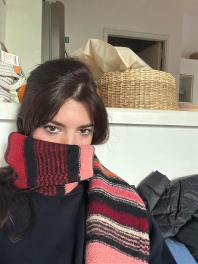 A disoriented close-up image shows a person with dark brown hair and brown eyes. The person is partially obscured by a red, black, and pink striped scarf or sweater. A wicker basket and beige cloth can be seen on the left side of the image, along with a box of "Candela" LED lighting. A black puffer jacket is visible in the upper right corner, along with a blue fabric item. The background appears to be a white wall. The image may be rotated or taken at an unusual angle. It is not possible to confirm the location as La Lucila, Argentina from the given image.