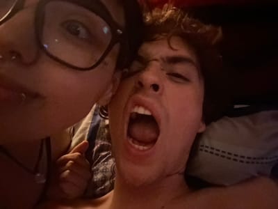 Two individuals lie side-by-side, viewed from an upside-down perspective. One person, possibly male, has their mouth wide open, appearing to be yelling or singing. The other, potentially female, wears glasses and looks towards the camera with a more subdued expression. The scene is dimly lit. One can observe elements like beddings and pillows, hinting at an intimate, personal setting.