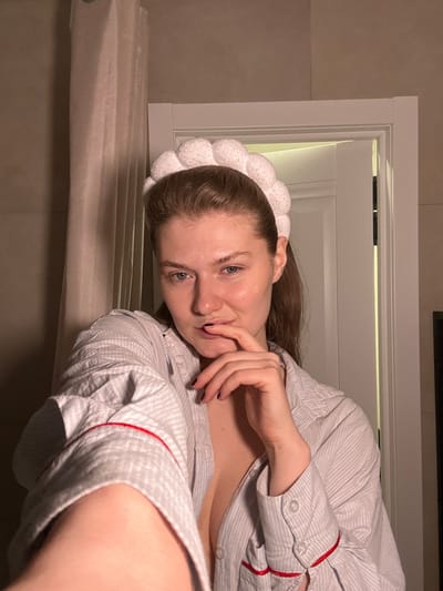 An indoor selfie captured in the city of Yanga-Yul, Russia, shows a female individual from the chest up. She has light skin, light-colored eyes, and brown hair styled back, secured by a white, textured headband. She wears a light-grey striped button-up shirt with red piping along the collar and cuffs, which is unbuttoned to reveal cleavage. Her left hand, with dark nail polish, is raised, with a finger touching her lower lip. She looks directly at the camera. The background includes a light-colored wall, a white curtain on the left, and a white door frame on the right, slightly ajar to a darker interior.