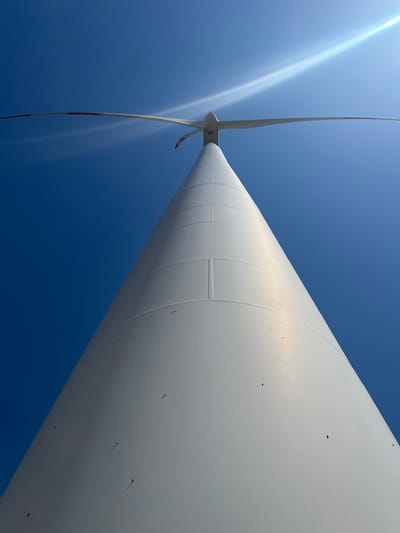 The image shows a close-up, low-angle view of a white wind turbine tower against a clear blue sky. The tower is segmented and tapers towards the top, where a large rotor with two blades is visible. The blades are white with red markings at the tips. A lens flare from the sun is prominent in the upper right quadrant of the image. The scene appears to be outdoors with no other objects or people visible. The location is identified as Rignano Garganico, Italy.