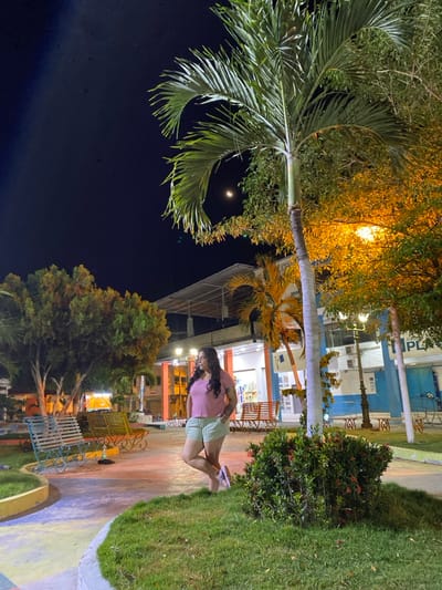 The image depicts a nighttime scene in a public square or park in General Villamil Playas, Ecuador. A dark sky with a visible crescent moon is overhead. In the midground, a woman with long, dark hair, wearing a light pink short-sleeved top and light green shorts, stands posing with her left leg crossed over her right, looking towards the upper left. Her left hand is in her pocket, and she is wearing light-colored sandals. She stands near the base of a prominent palm tree. To her left, paved pathways are visible with multi-colored sections (yellow, blue), along with several park benches (light blue/green and yellow/orange) and other trees. To her right, another tree with orange-illuminated foliage and a streetlight are present. In the background, illuminated multi-story buildings are visible, some with white, orange, and blue facades. One building partially displays the word "PLAYAS". Artificial lighting casts warm orange hues on some elements and cooler light on others, illuminating the scene.