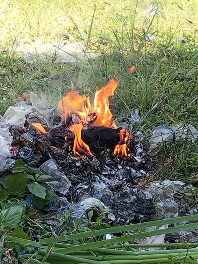 A fire blazes amidst scattered trash in a grassy area. Flames lick upwards from a pile of dark ash and burning debris, including plastic bottles and bags. Smoke rises into the air. The surrounding vegetation consists of weeds and grass. Some of the plastic debris is visible beyond the fire, discarded within the greenery.