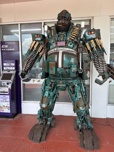 A large, life-sized statue of a robot made from scrap metal stands on a red tiled floor in front of a building with a glass facade. The robot is predominantly green with rust accents and features intricate mechanical details, including what appear to be machine gun barrels on its arms and a belt of bullets across its chest. It has a beard and blue eyes. To the left of the robot is a purple ATM machine with Thai writing and logos for various card types. The building behind the robot appears to be a commercial establishment, possibly a convenience store, with a "7-Eleven" logo visible through the glass. The overall environment suggests an outdoor urban setting, likely in a city like Tha Ruea, Thailand, given the Thai text on the ATM.