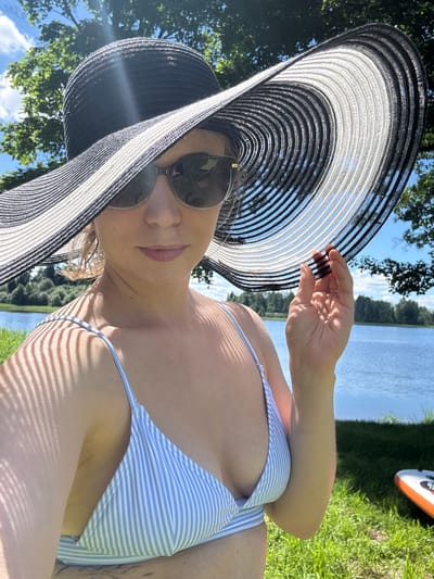 A woman is taking a selfie outdoors by a lake. She is wearing a wide-brimmed black and white striped hat and sunglasses. Her bikini top is blue and white striped. Her right hand is raised, touching the brim of her hat. The background shows a body of water, green grass, trees, and a blue sky with clouds. A paddleboard is visible on the grass to the right. The woman appears to be in Lendži, Latvia.