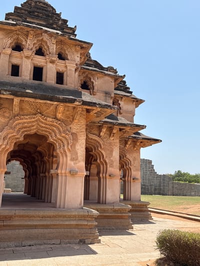This outdoor, medium-angle shot features the Lotus Mahal building located in Kamalapura, India. The building is constructed from a tan-colored stone. The perspective is looking up at the side and roof of the building. The roof has a series of triangular elements that are stacked on top of each other. The side of the building features a repetitive archway design. The arches are intricately carved with floral and geometric designs. The building has windows. A stone wall with stone stairs on the top right part of the frame. Above the wall is grass, trees, and blue sky. The light is bright and diffused.