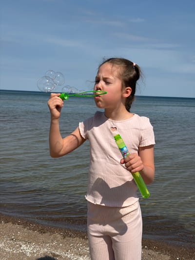 A young girl is blowing bubbles on a beach in Plieņciems, Latvia. She is wearing a light pink, striped, short-sleeved shirt and matching pants. Her hair is pulled back into a ponytail with a pink hair tie. She is holding a green bubble wand and blowing into it, creating a cluster of bubbles. The background shows the beach with sand and water, and a clear blue sky. The scene is well-lit.