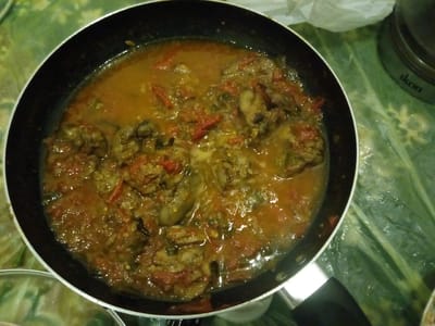 A black frying pan containing a saucy stew of meat and tomatoes is placed on a green, patterned surface. To the right of the pan, a silver canister with the word "ikon" printed on it is visible. A white, crinkled object is behind the canister. The scene appears to be indoors, potentially in a kitchen. There are no people visible in the image. The lighting is dim, and the image is taken from a top-down perspective.