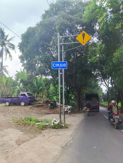 A scene on a paved road in Kecamatan Cisarua, Indonesia, with a blue sign reading "Cimahi" mounted to a metal pole. The pole also supports a yellow diamond-shaped sign with a black exclamation mark. The scene is viewed from the perspective of a person moving down the road. A person is riding a motorcycle in the middle of the road, with another person riding a motorcycle in the distance. A dark green truck is driving towards the right edge of the road. A purple pickup truck is parked on the left side of the road. There is a grassy area on the left side of the road and trees surrounding the area.