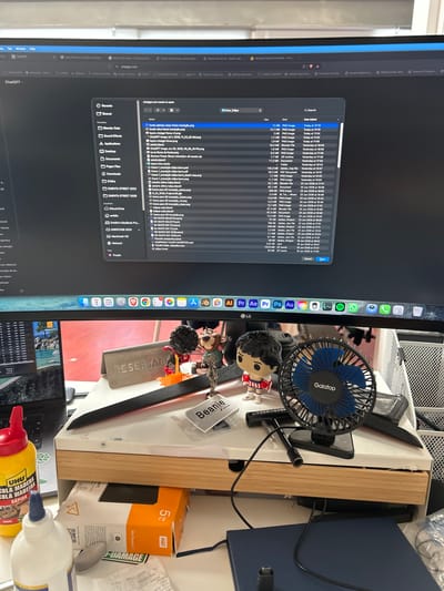 The image shows a desk with a computer monitor displaying a file explorer window, likely on a macOS system, with a list of files. The file names suggest a project involving 3D transitions, Blender files, and possibly image editing, with dates ranging from June 2026. Among the files are images with names like "fundo plantas roxas frame transição.png" and "fundo relva frame transição.png", indicating a visual or artistic project.
On the desk, to the right of the monitor, are various objects. A small blue and black USB fan labeled "Gaiatop" is visible, along with two small figurine toys. There's also a black knife, a wooden stand, a dark blue folder or book, and a white box labeled "WD My Passport" with "5 TB" indicated. A bottle of "UHU COLA MADERA RÁPIDA" (wood glue) is also prominent. A receipt with text in Portuguese is partially visible. The overall setting appears to be a home or office workspace, possibly in Cacém e São Marcos, Portugal, given the context. The lighting suggests it's daytime.