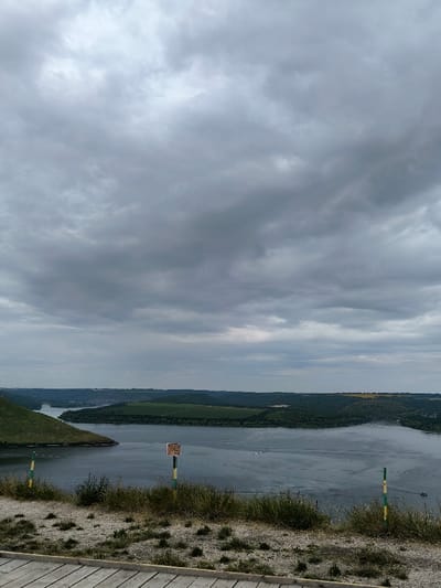The image displays an elevated panoramic landscape view of the Dniester River canyon, located near the village of Horaivka, Khmelnytskyi Oblast, Ukraine. The scene is observed under a completely overcast sky filled with dense, uniform grey clouds.
In the immediate foreground, the bottom left shows a portion of a light-colored wooden plank surface, consistent with a boardwalk or viewing platform. The ground directly in front of the viewpoint is composed of light-grey, dry soil or gravel with scattered patches of green grass and weeds. Three slender poles are visible: one partially obscured on the far left with green and yellow stripes, a central weathered wooden post bearing a rectangular, light-brown sign with dark text, and another green and yellow striped pole on the far right, partially obscured by vegetation.
The midground is dominated by the wide, calm expanse of the Dniester River, which flows from the left of the frame and curves towards the right. The river's surface exhibits subtle ripples and what appear to be distant wakes from a small, unidentified watercraft, barely visible in the upper right quadrant of the water body. The river is bordered by steep, verdant hills and densely forested areas on the opposite bank.
The background extends into a series of undulating hills and plateaus, covered with a mosaic of dark green foliage and lighter green or yellowish agricultural fields. The distant horizon merges indistinctly with the low cloud base. No individuals or discernable structures other than the poles and implied platform are present in the immediate vicinity.