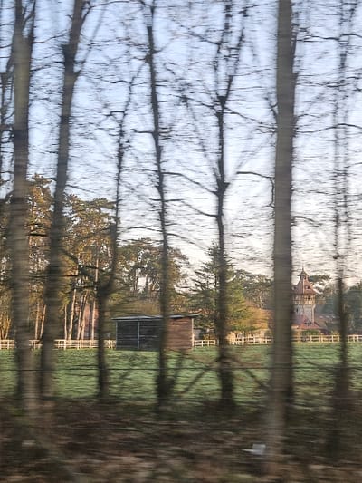 The image depicts a rural landscape near Émancé, France, captured with significant vertical motion blur. In the immediate foreground, numerous bare deciduous trees appear as vertical streaks, obscuring parts of the scene. The midground features a vibrant green field, likely pastureland, bordered by a white, multi-rail fence that spans horizontally across the frame. Centered within this field is a dark, rectangular wooden shed with a sloped roof and an open front section. Further in the background, to the right, stands a distinctive building, identified as the Château d'Émancé, characterized by a prominent multi-story tower with a conical roof, visible above the treeline. Surrounding the château and extending across the background are dense clusters of trees, some evergreen and others appearing autumnal or bare, under a pale blue sky. No individuals or specific actions are discernible.