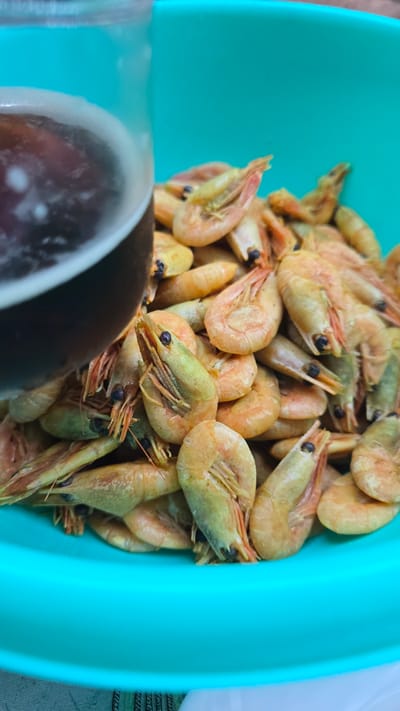 A close-up shot shows a bright teal bowl filled with cooked shrimp. The shrimp are a pale orange-pink with some greenish hues, and their antennae and legs are visible. Many of the shrimp have their heads pointed upwards, and their eyes are dark. The bowl is positioned at an angle, with a portion of a dark, glossy serving utensil or bowl edge visible in the lower left corner of the frame. The background is out of focus, but appears to be a patterned surface, possibly a tablecloth or placemat, and a hint of a light-colored surface. The lighting suggests daytime, and the overall impression is one of a prepared dish ready to be served. There is no visible text in the image.