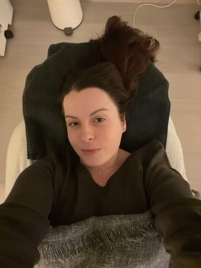 The scene is an indoor room located in Callbach, Germany. A single female individual is depicted lying supine on a white upholstered examination or treatment table. Her head rests on a dark gray waffle-knit towel or blanket, with her dark brown hair spread above it. She is wearing a dark-colored, long-sleeved V-neck top, a delicate gold-toned necklace with a small crescent-shaped pendant, and small stud earrings. Her eyes are open, directed towards the camera, and her lips display a subtle smile. Her lower torso is covered by a light gray woven blanket with fringed edges. The floor is a light-colored hard surface. In the upper right background, a white cylindrical pedal bin with a dark base is visible, alongside the corner of a white cabinet or medical cart with a dark wheel. A thin white cable traverses the floor near these objects. Lighting is even and ambient.