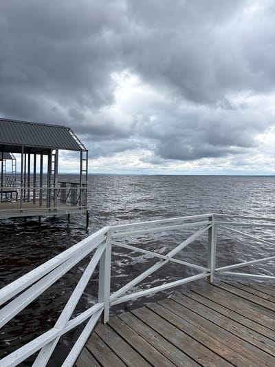 The image depicts a pier on a body of water under a cloudy sky. The pier is constructed of wooden planks and has a white metal railing. To the left, there is a structure with a metal roof supported by metal poles, appearing to be a covered area or boathouse. The water is dark and choppy, with visible ripples. In the distance, a faint horizon line separates the water from the sky. The sky is filled with heavy, dark gray clouds, suggesting overcast or stormy weather. There are no people or animals visible in the scene. The location is specified as Kostovaty, Russia.