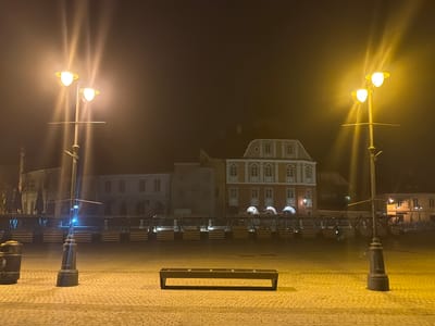The image displays the Piața Mare (Large Square) in Sibiu, Romania, at night, under conditions of significant atmospheric haze or fog. Two ornamental streetlights, each featuring two upward-facing yellow lamps, frame the foreground on the left and right, their light creating vertical streaks due to the atmospheric diffusion. The ground is paved with light-colored cobblestones. Centrally located in the mid-foreground is a dark, rectangular public bench with a slatted top. To the left, near the base of the left streetlight, a dark grey public waste bin is visible. Across the midground, a continuous line of elevated structures, consistent with seasonal market stalls or outdoor dining terraces, extends horizontally. These structures have wooden bases, individual steps, and some feature translucent dome-shaped coverings. In the background, behind these structures, several multi-story historic buildings are visible. The most prominent is a brick-red building with multiple white-framed windows and an ornate roofline featuring dormers. The scene is devoid of any individuals.