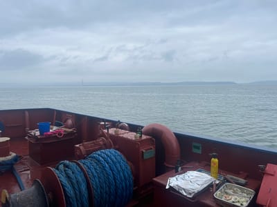 The image shows the deck of a maroon-colored vessel at sea on an overcast day, likely in Belfast Lough, near Bangor, Northern Ireland. In the immediate foreground, a large spool of coiled blue rope is prominent on the wet deck. To its left, another smaller spool with dark rope is visible. Further back on the port side, a blue bucket, a pink rag, and various pieces of industrial machinery, including an electric motor and pipework, are situated on a raised platform. A "NO STEP" sign is affixed to one of the red structures. On the starboard side, an open metal tray containing what appears to be raw seafood (possibly mussels or scallops with onions) and a rectangular container wrapped in aluminum foil rest on a flat surface of the deck machinery. Adjacent to these items are a yellow gas canister with an attached torch head, a dark bottle, and a pair of tongs. The water in the mid-ground is calm and gray, reflecting the cloudy sky. The distant horizon reveals a hazy shoreline with low hills and distinct industrial features, including a tall, slender chimney-like structure. No individuals are visible in the scene.