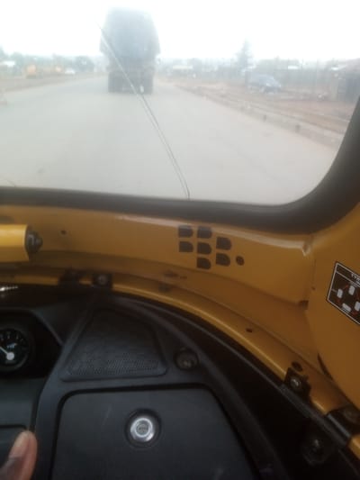 The image appears to be taken from inside a yellow vehicle, possibly a three-wheeled auto-rickshaw or tuk-tuk, looking out through the windshield. The foreground shows a portion of the vehicle's interior, including a black dashboard with a circular gauge and a black textured panel with a silver circular emblem. A hand is visible on the left side, partially obscuring the view.
The windshield is slightly smudged and has two thin, dark lines running across it, possibly from debris or damage. Through the windshield, a road is visible with a large, dark truck in the distance, partially obscured by haze. Other vehicles and structures are faintly visible in the background, suggesting an outdoor, possibly urban or semi-urban setting. The road appears to be paved, and there are some barriers or poles along the side. The sky is overcast, indicating a cloudy day.
On the yellow bodywork of the vehicle, to the right of the windshield, there is a black decal that appears to read "BBB." Further to the right, there is a black sticker with white text that includes the words "TYRE" and "FRONT." The scene suggests the vehicle is in motion or stopped on a road in Tundun Wada South, Nigeria. The overall mood is somewhat hazy and subdued, possibly due to the weather conditions.