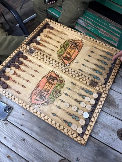 A wooden backgammon board is laid out on a wooden table. The backgammon board is set up with checkers and dice. The board is decorated with a green, blue and red pattern. The board is sitting on a table outside. The table is made of wooden planks, and there is a green wooden bench in the background. The image was taken in Mazkeret Batya, Israel. The coordinates (latitude: 31.833, longitude: 34.82) suggest the image was taken near the Mazkeret Batya cemetery. The image was taken in a public place, most likely a park. The image is taken from a high angle, as if the photographer is looking down at the board. It is likely that someone is playing backgammon. The scene appears peaceful and casual. The image is well-lit, and the colors are vibrant.