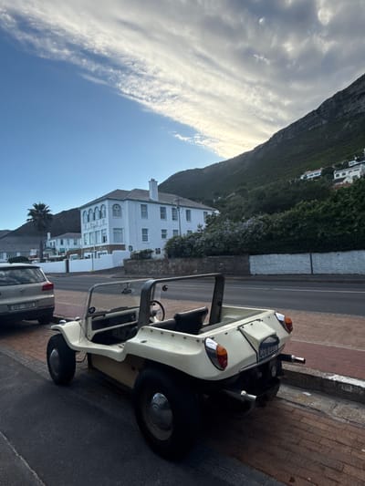 The image shows a white-colored, open-top, vehicle parked on a paved road in Muizenberg, South Africa. A silver-colored car is parked behind it. A large white building with multiple windows, likely a hotel or apartment complex, is visible across the street. A mountainside covered with vegetation forms the backdrop. The sky is partly cloudy.