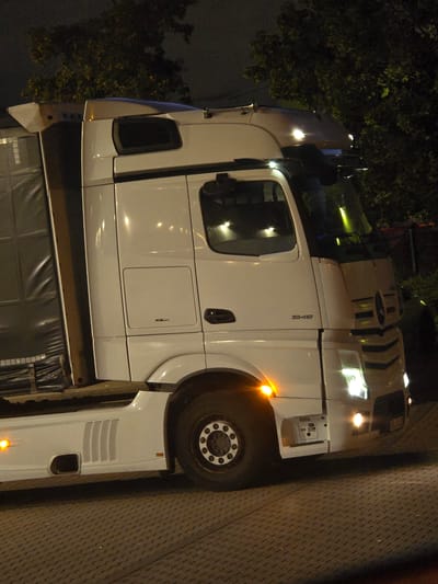 A white Mercedes-Benz Actros 1848 semi-truck is parked on a patterned brick surface at night. The truck has a large white cab with tinted windows, chrome accents on the grille, and visible headlights and turn signals. The truck's trailer is partially visible on the left, covered with a dark grey tarp. Trees with yellow and green foliage are visible in the background, illuminated by artificial light. The scene is likely in Zeppelinheim, Germany, given the context.
