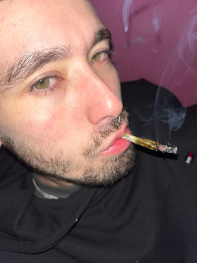 A close-up, tilted shot shows a person with light skin, stubble, and green eyes holding a lit joint in their mouth. The person has a silver hoop nose piercing. Smoke rises from the joint. The background is partially pink and partially dark, with what appears to be a Tommy Hilfiger logo visible near the dark area. The location is not discernible from the image provided.