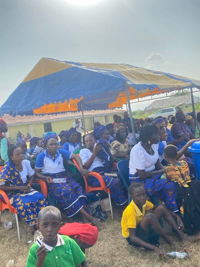 The image depicts an outdoor gathering in Itobe, Nigeria, under a blue and orange striped canopy tent on a sunny day. Numerous individuals, primarily adults and children, are present. Many adult women occupy orange and blue plastic chairs, dressed in matching blue patterned skirts and head wraps, often paired with white blouses or blue polo shirts. Some individuals, including children, are seated directly on the dry, grassy ground.
In the foreground, a young boy in a green collared shirt with a beaded necklace looks towards the viewer, holding an object to his mouth. To his right, another child in a yellow polo shirt sits on the ground. Various plastic water bottles are visible on the ground and held by attendees. In the mid-ground, a young child with a multicolored dress is held by an adult. A blue plastic drum or bin is situated to the right.
The background reveals single-story buildings with light-colored walls, possibly yellow or off-white, and dark roofs. Beyond the structures, a white SUV or truck is parked, and sparse vegetation is visible under a clear, bright sky. The sun is high in the frame, casting strong light.