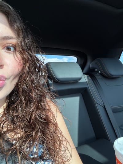 A selfie in a car shows a young woman with wet, curly brown hair and brown eyes. Her lips are slightly pursed. The car's interior is visible, featuring black leather seats with light stitching and a safety belt. A window provides a glimpse of the sky with light clouds. A drink container is visible in the top right corner.