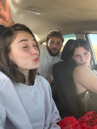 Three individuals are depicted inside a vehicle, located within the Ramat Negev Regional Council, Israel. In the foreground, positioned to the left, is a female with dark brown hair, wearing a light grey long-sleeved sweatshirt. Her eyes are closed, and her lips are pursed in a kissing gesture. Above her head, a human hand with a light skin tone is partially visible, forming a V-sign. In the middle ground, centrally positioned, is a male with dark curly hair and a beard, wearing a light-colored collared jacket. He is looking directly at the camera with a neutral expression. To the right, another female with dark brown hair is visible, wearing a dark top with cut-out patterns on the side. Her lips are also pursed in a kissing gesture, and her head is slightly tilted. The interior of the vehicle features a light-colored headliner and dark seatbacks. A crumpled red plastic bag is visible in the lower right corner of the frame. Through the vehicle's windows, an exterior environment consistent with an arid or desert landscape is discernible, characterized by light-colored ground and a bright sky.
